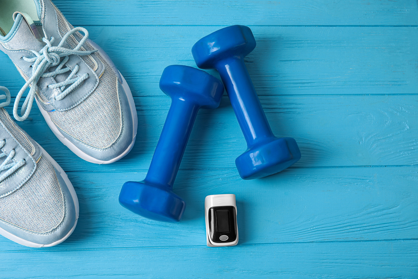 Pulse Oximeter, Dumbbells and Sport Shoes on Color Wooden Background, Closeup